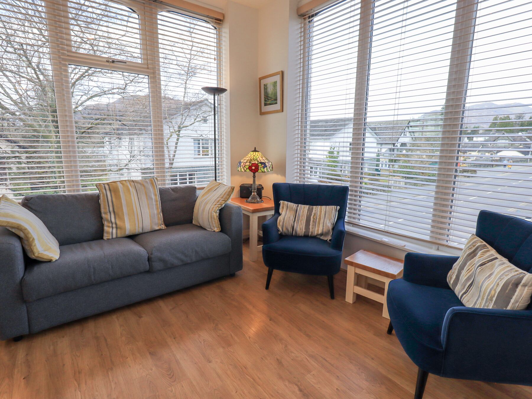 Een woonkamer met een grijze bank, een blauwe fauteuil en een kleine tafel met een lamp.