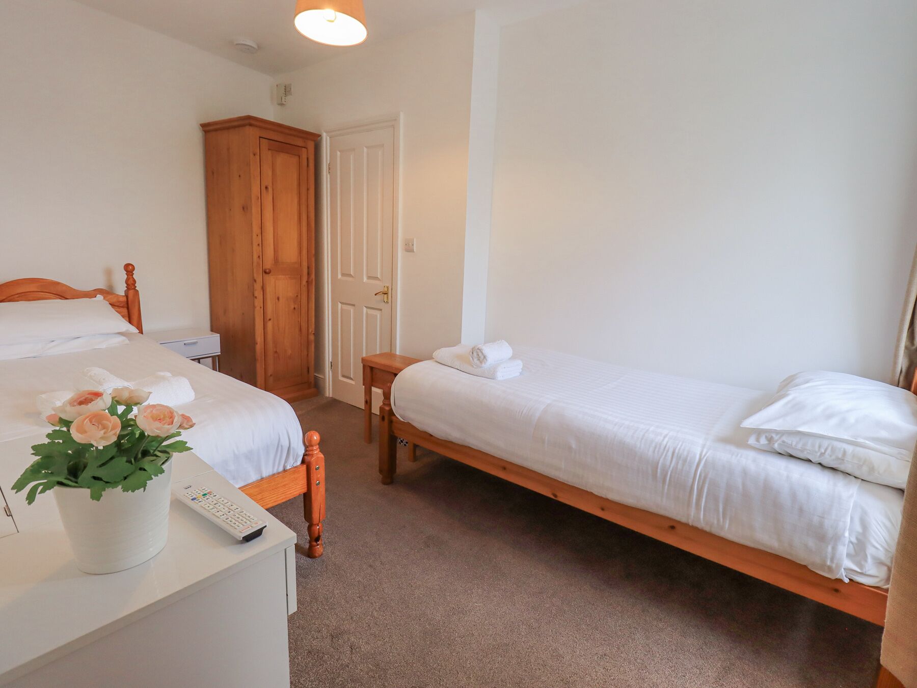A bedroom with two single beds, a wooden wardrobe, a bedside table with a remote control, and a potted plant.