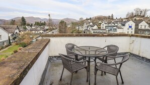 A balcony with wicker chairs and a table, overlooking a residential area with houses and mountains in the background. - Windows (Ambleside)