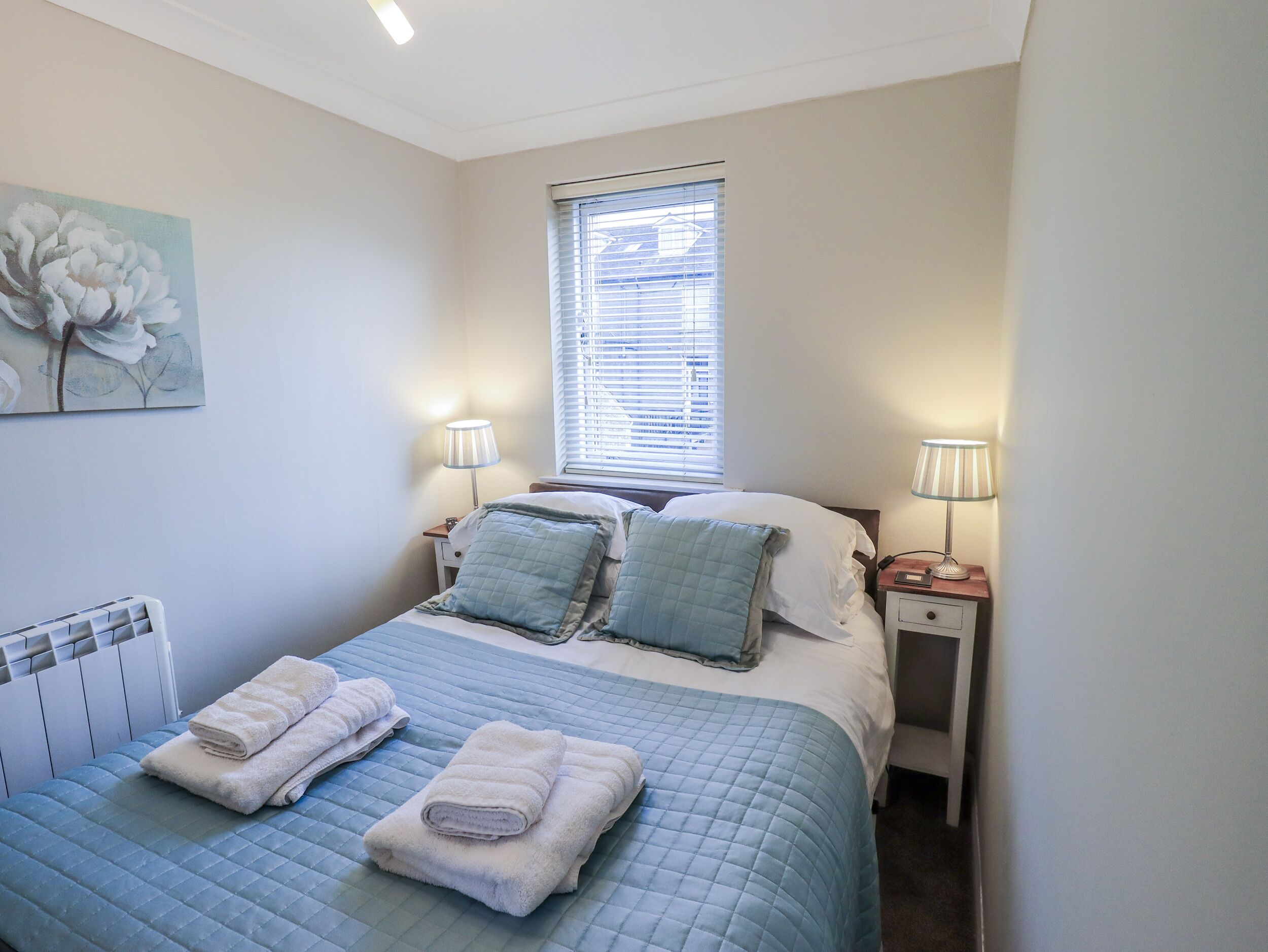 A bedroom with a bed, two bedside tables, a radiator, and a window with blinds.