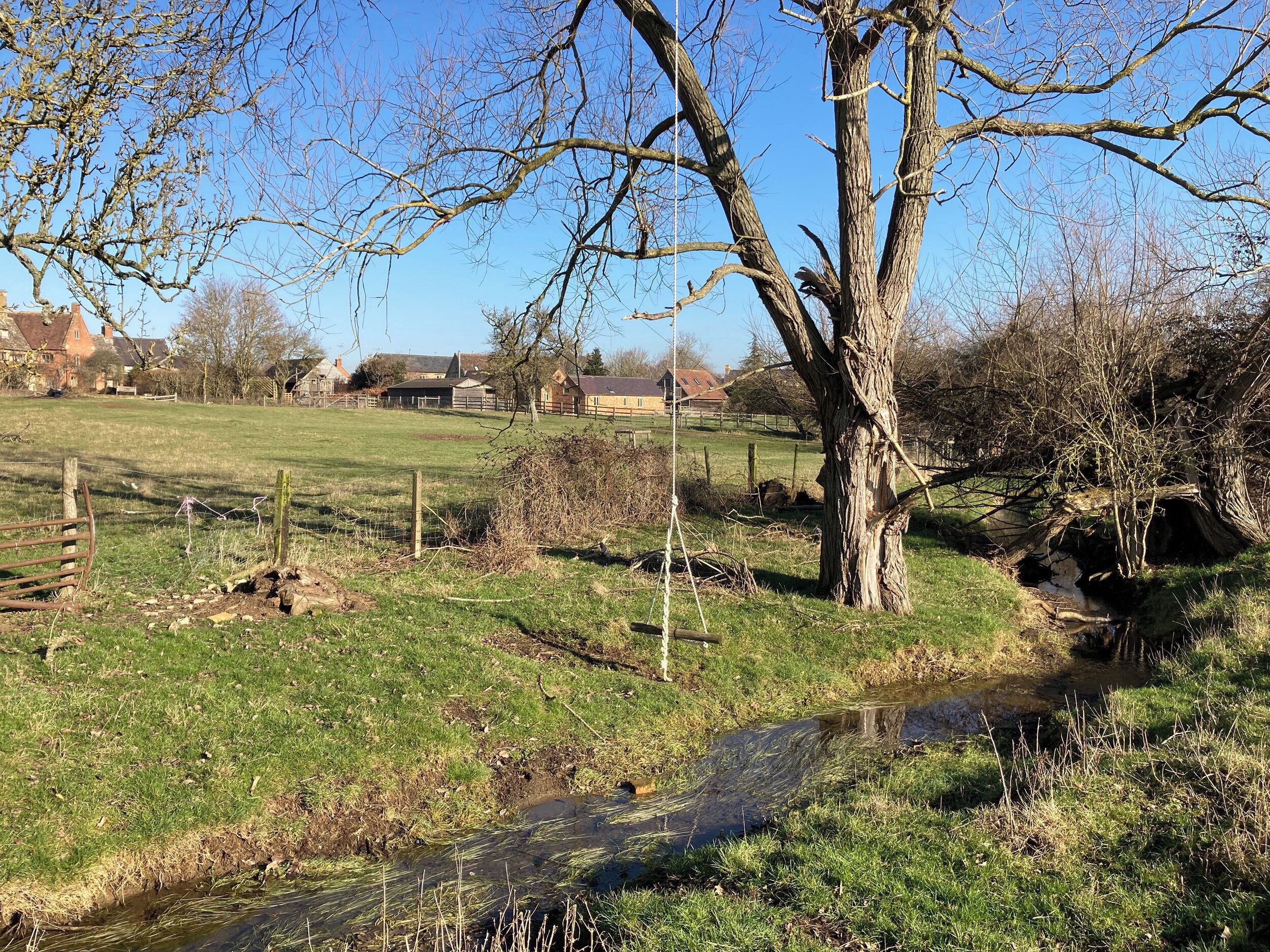 Un paisaje rural con un campo cubierto de hierba, un pequeño arroyo y árboles desprovistos de hojas.