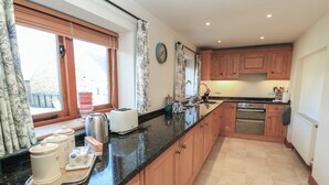 Une cuisine avec des meubles de rangement en bois, un plan de travail noir, une fenêtre avec des rideaux, un grille-pain, une bouilloire et une horloge au mur.