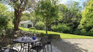 Une terrasse avec une table et des chaises, entourée d’un jardin et d’un petit bâtiment.