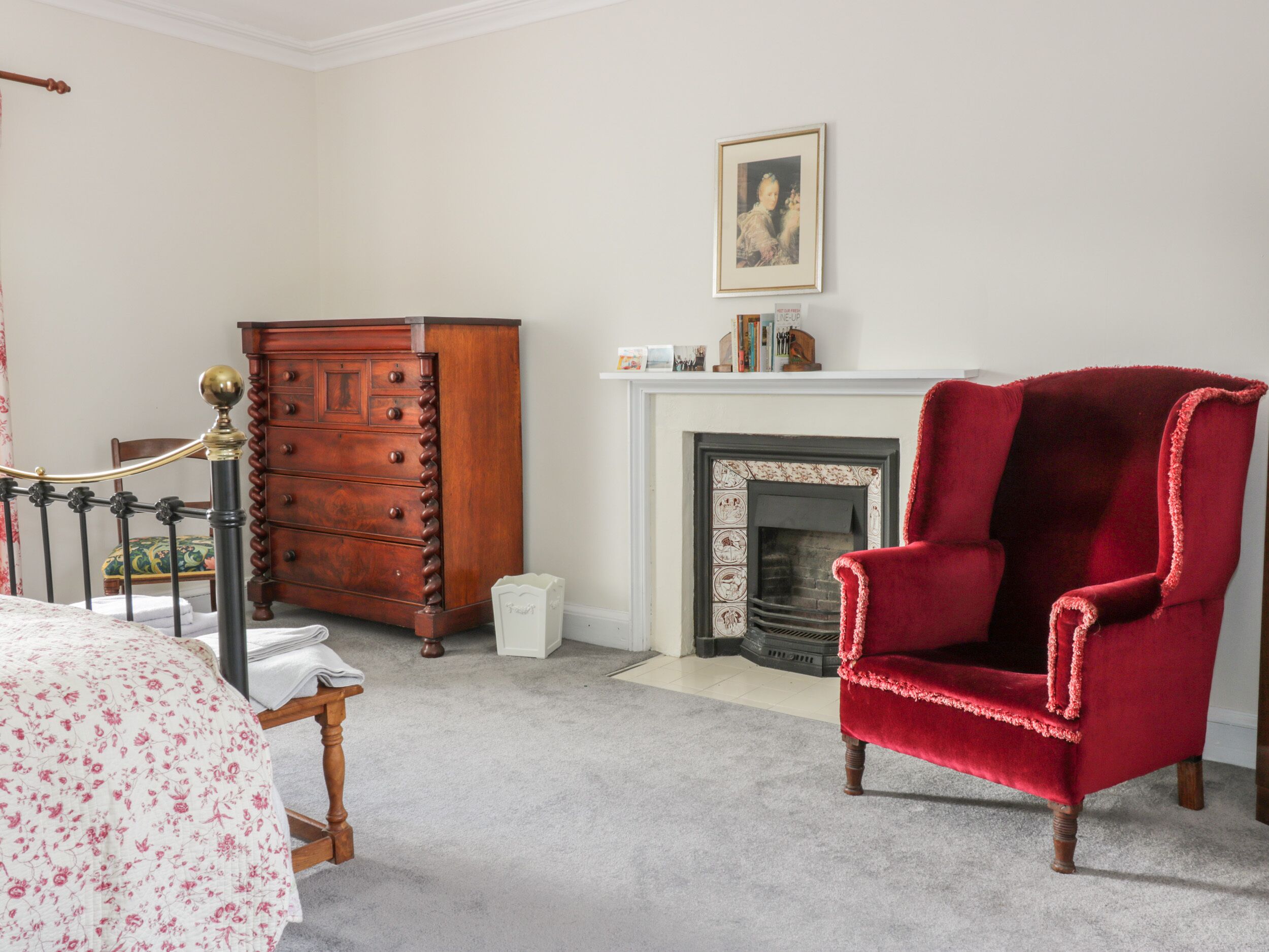 A bedroom with a wooden dresser, a red armchair, a bed with a floral bedspread, and a fireplace.
