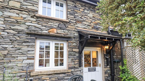 A stone cottage with a white door, a window, and a small outdoor seating area with a table and chairs. - Cobblestone Cottage (Ambleside)