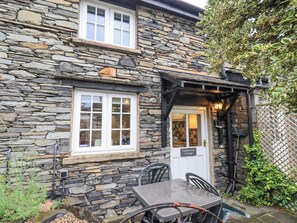 A stone cottage with a white door, a window, and a small outdoor seating area with a table and chairs.