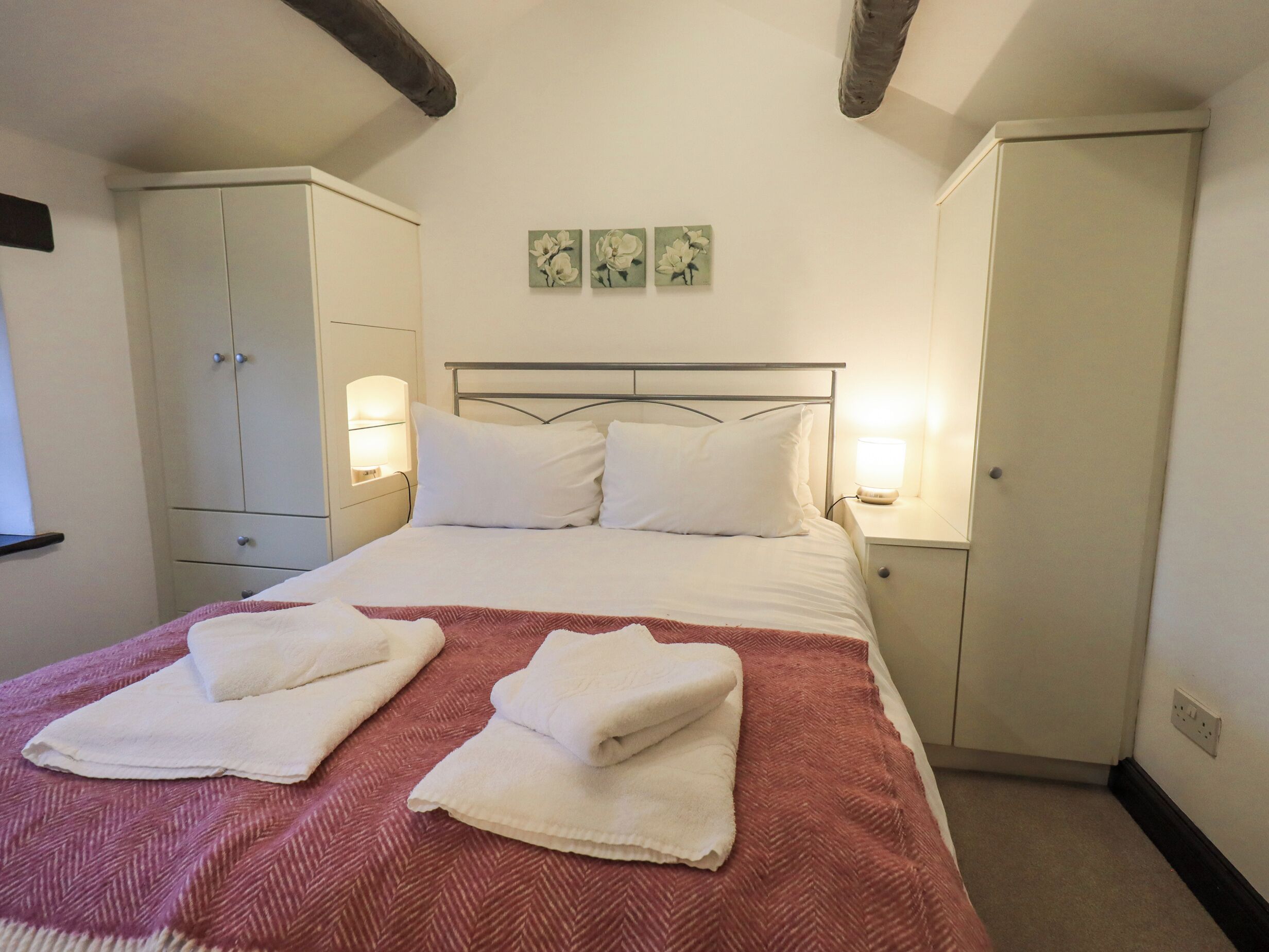 A neatly made bed with white linens and two folded towels, a wooden ceiling beam, a white wardrobe, and a nightstand with a lamp.