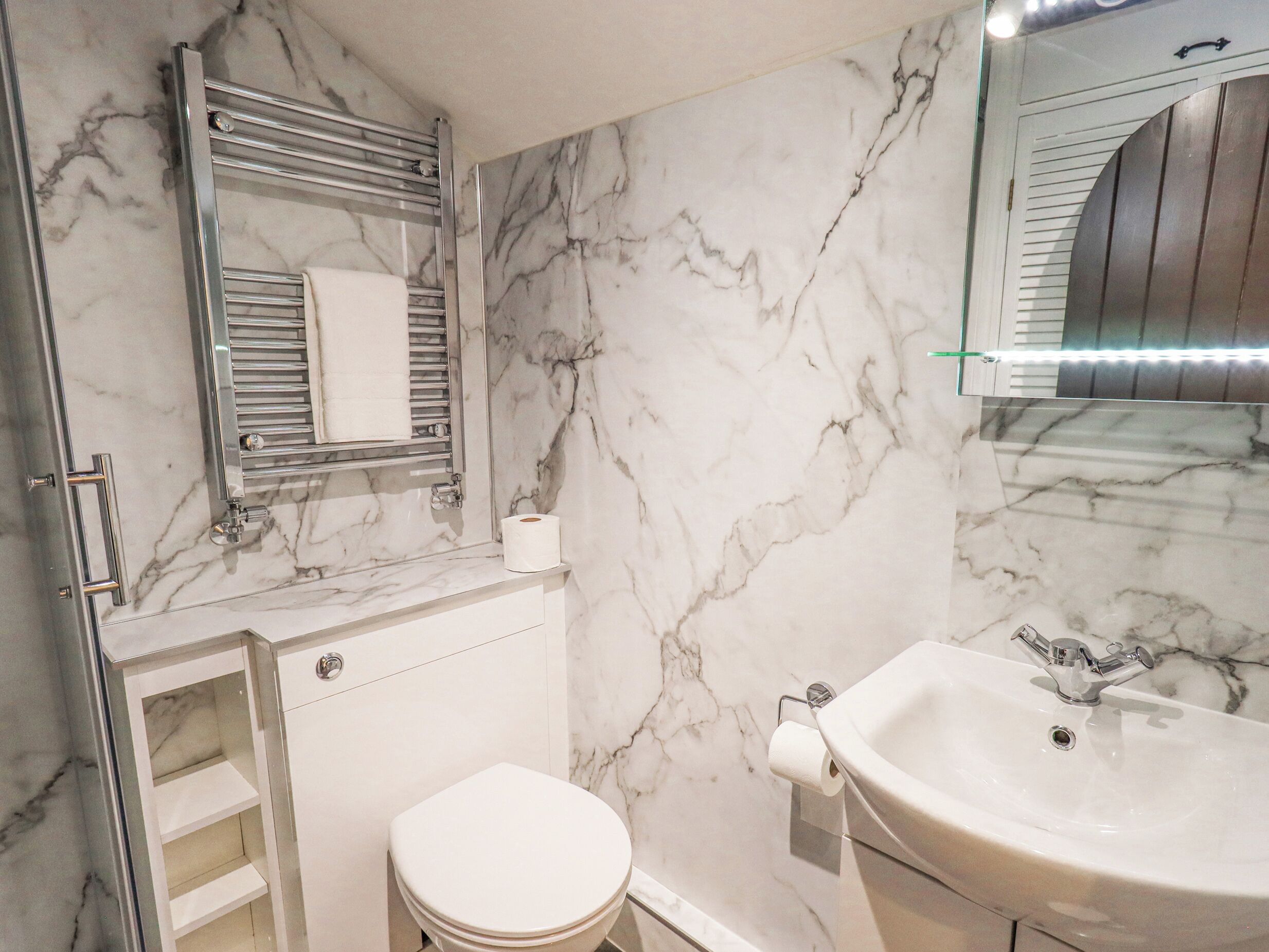 A bathroom with a marble wall, a toilet, a sink with a mirror above it, and a towel rack.