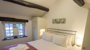 A neatly made bed with white linens and a pink blanket, a bedside lamp, and a window with a view of the outside. - Cobblestone Cottage (Ambleside)