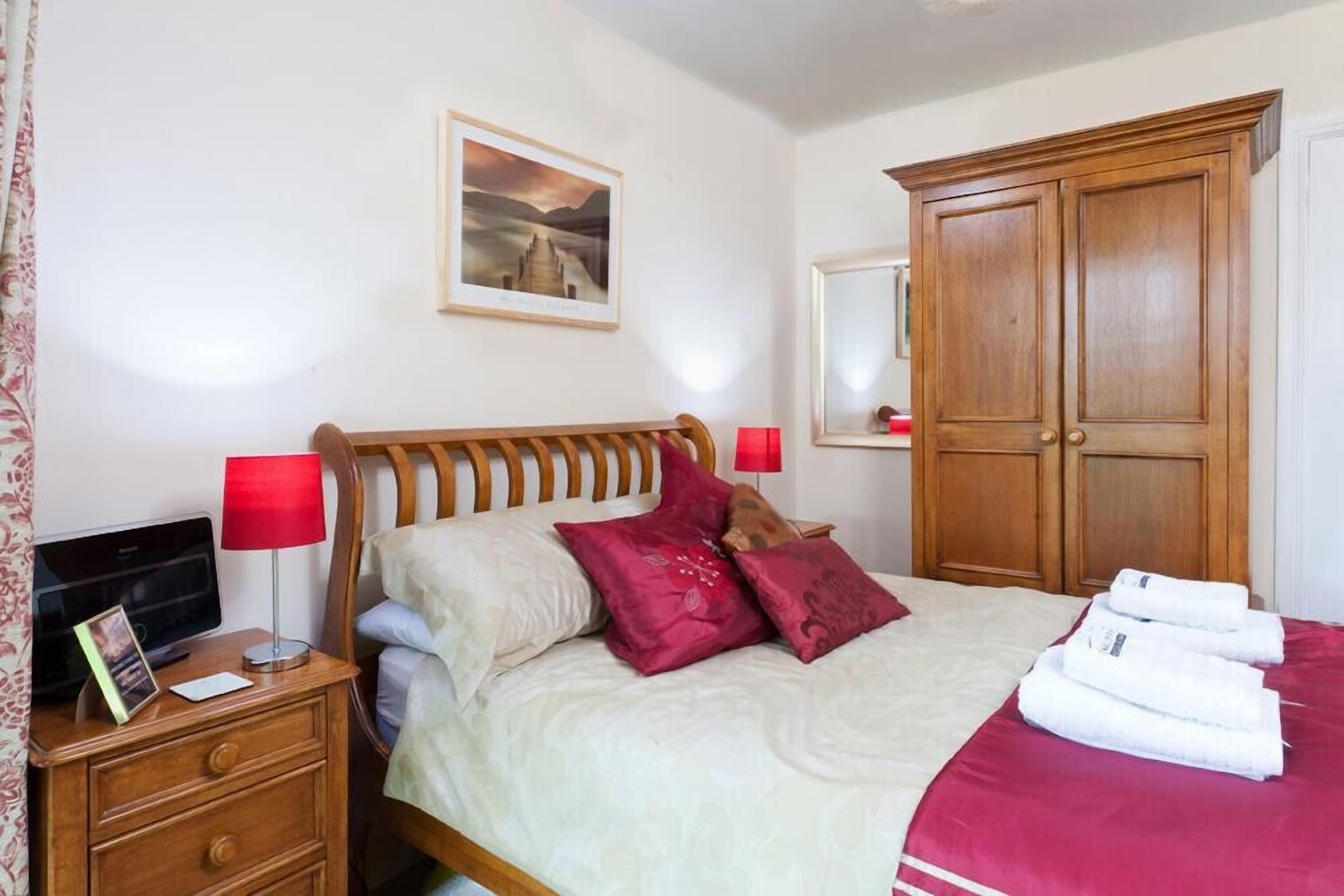 A neatly made bed with a red blanket, a wooden nightstand with a TV and a red lamp, and a large wooden wardrobe.