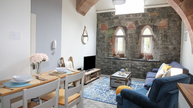 Uma sala de estar acolhedora com uma parede de pedra, uma mesa de jantar de madeira com cadeiras, um sofá azul e uma televisão.