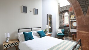 Quarto com cama, mesa de cabeceira com abajur, espelho e pequena mesa com um vaso de flores.