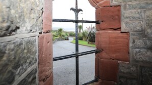 Visão através de um arco de tijolos com portão de metal, conduzindo a uma área pavimentada com vegetação e edifícios ao longe.