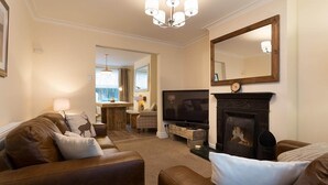 A living room with a fireplace, a television, and a dining area with natural light. - Noble Cottage (Keswick)