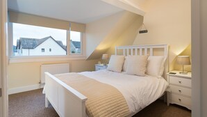 A bedroom with a bed, bedside tables, a window, and a view of a house outside. - Noble Cottage (Keswick)