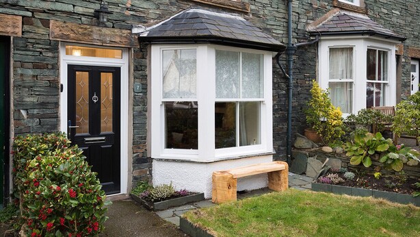 A stone house with a black door, a bay window, and a wooden bench. - Noble Cottage (Keswick)