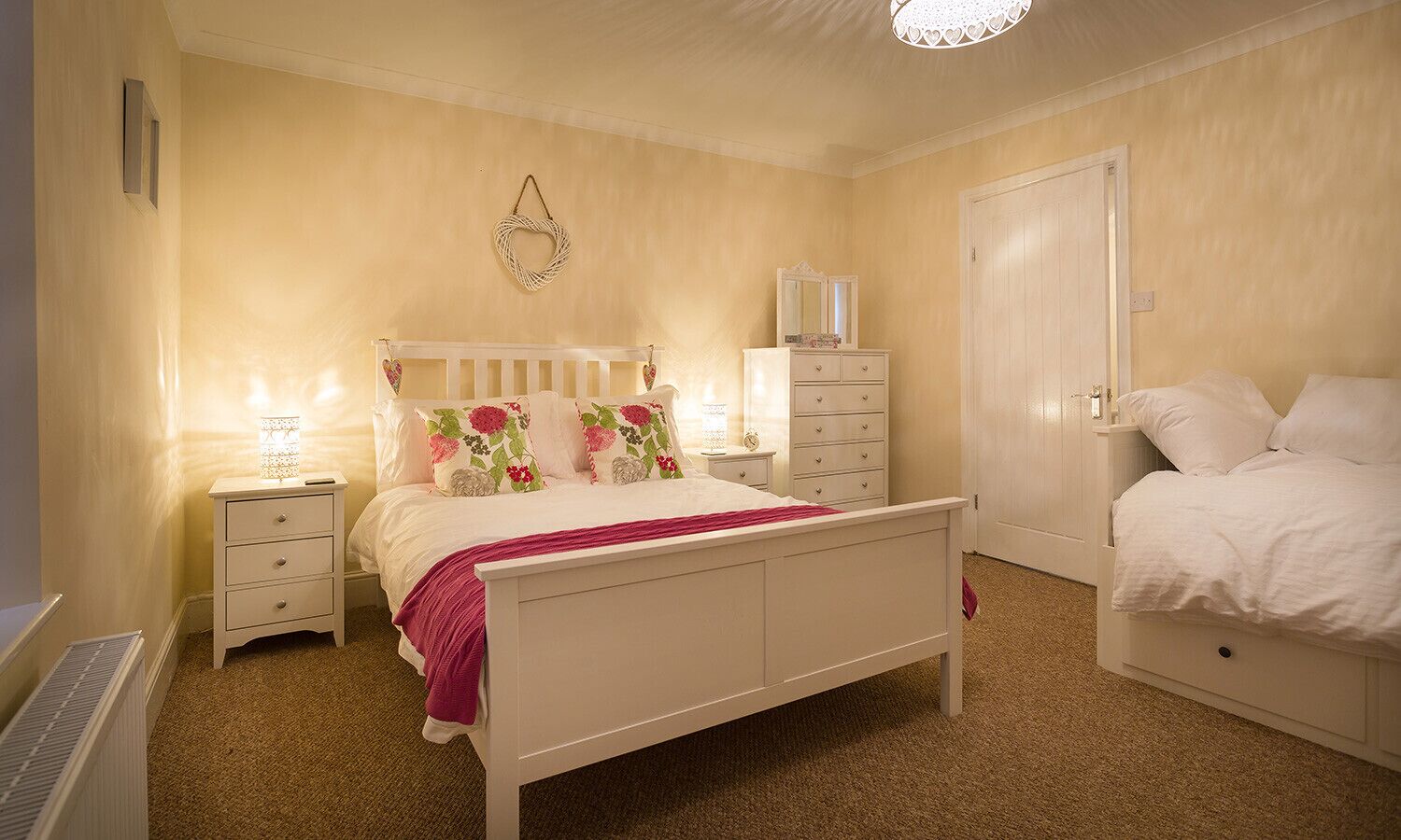 Quarto com cama branca, mesa de cabeceira com abajur, cômoda e porta.