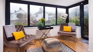 A modern living room with two brown sofas, a patterned ottoman, a small table, and a large window with a view of houses and greenery. - Foxrock (Windermere)