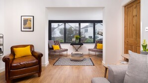 A modern living room with a brown leather sofa, two armchairs with yellow cushions, a patterned ottoman, and a large window offering a view of the outside. - Foxrock (Windermere)