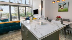 A modern kitchen with a marble island, large windows, and a dining area with a blue sofa. - The Hay Barn (Houghton le Spring)