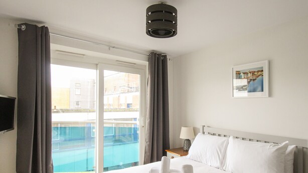 A hotel room with a bed, a nightstand, a lamp, a painting, and a view of a swimming pool. - Nautical Breeze (Weymouth)