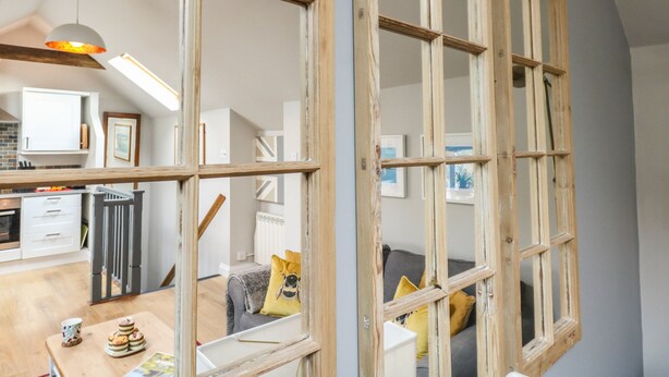A room with a large wooden-framed window, a kitchen area with white cabinets, and a dining table with a cup and pastries. - Little Brook (Weymouth)