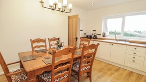 A dining area with a wooden table and chairs, a chandelier, and a kitchen with white cabinets and a window. - Hardy Cottage (Tyn-y-Gongl)