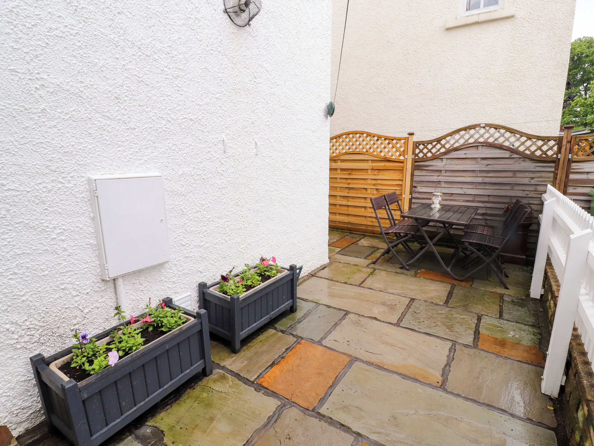 A patio area with stone flooring, two planters with flowers, a wooden fence, and a table with chairs.