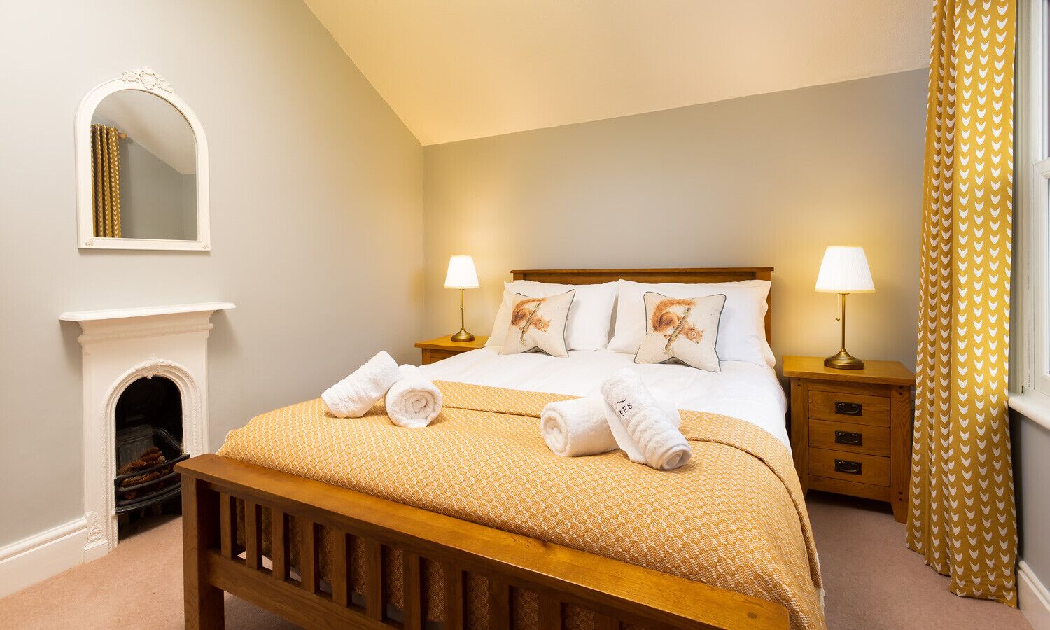 Quarto com cama de madeira, duas mesas de cabeceira com abajures, lareira e janela com cortinas amarelas.