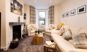 A cozy living room with a fireplace, two sofas, a coffee table with books and a wine glass, and a view of a vehicle outside. - Dovedale (Keswick)