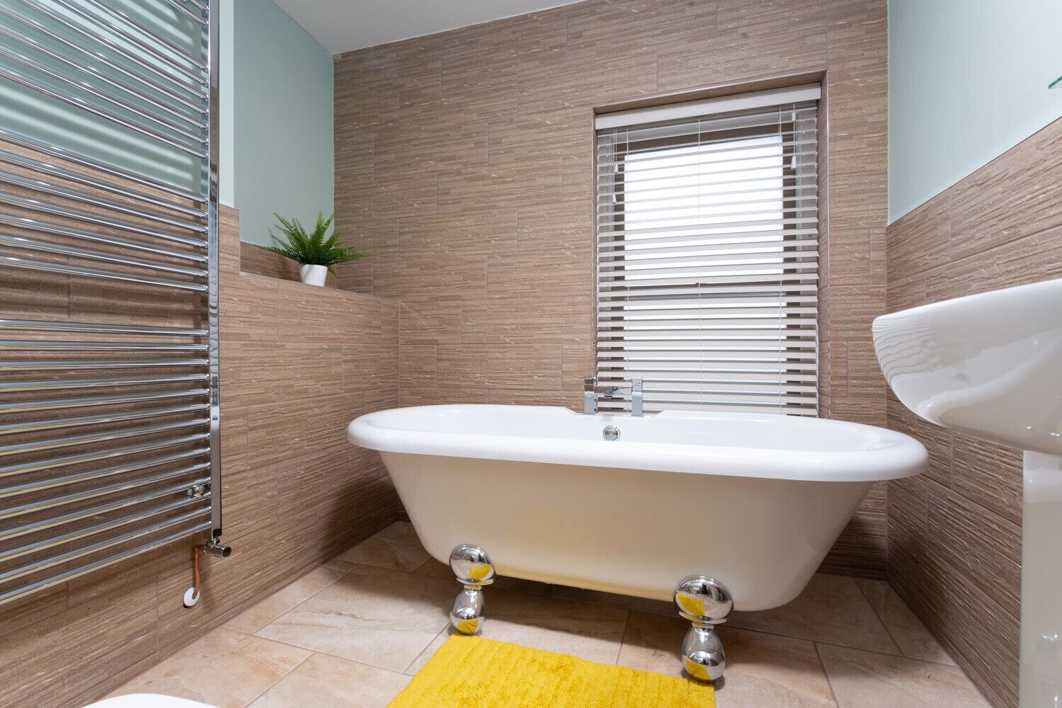 A bathroom with a freestanding bathtub, a window with blinds, a sink, and a yellow bath mat.