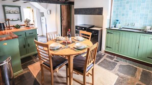 On aperçoit un coin repas avec une table en bois dressée pour quatre personnes, entourée de chaises. Au fond, on distingue une cuisine avec des meubles verts et un dosseret carrelé.