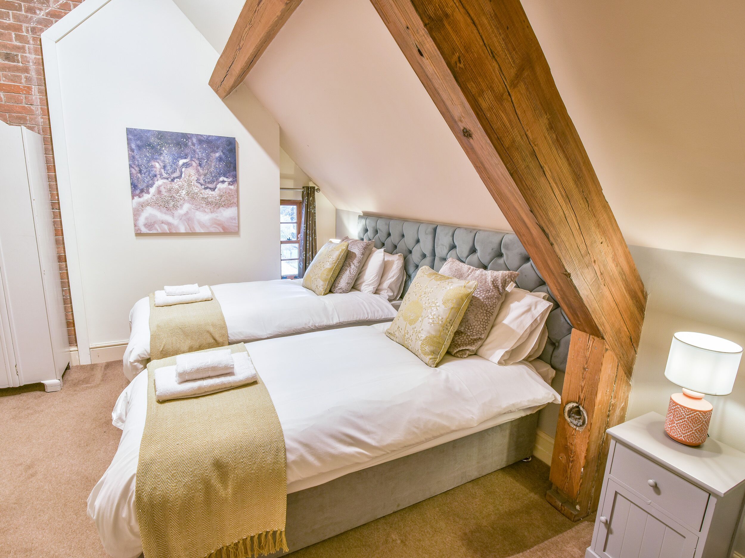 A bedroom with two beds, a wooden ceiling beam, a nightstand with a lamp, and a painting on the wall.