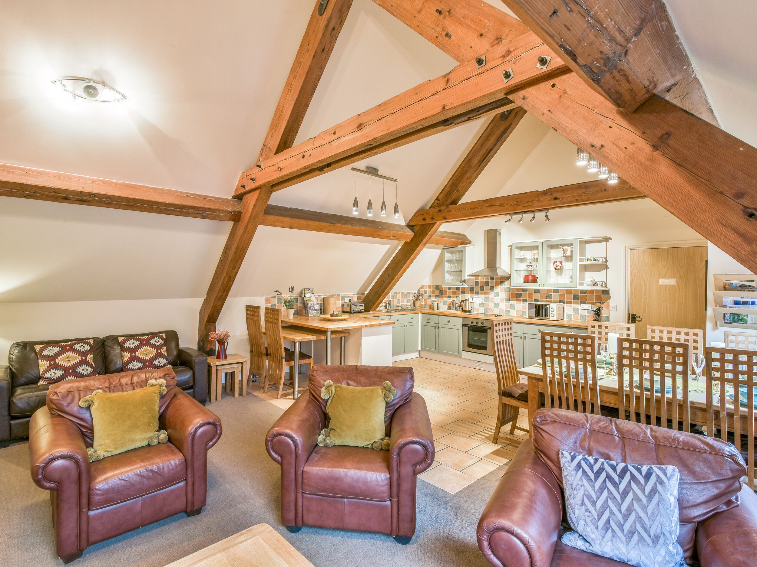 A spacious loft with exposed wooden beams, a cozy seating area with leather sofas, a kitchen with modern appliances, and a dining area.