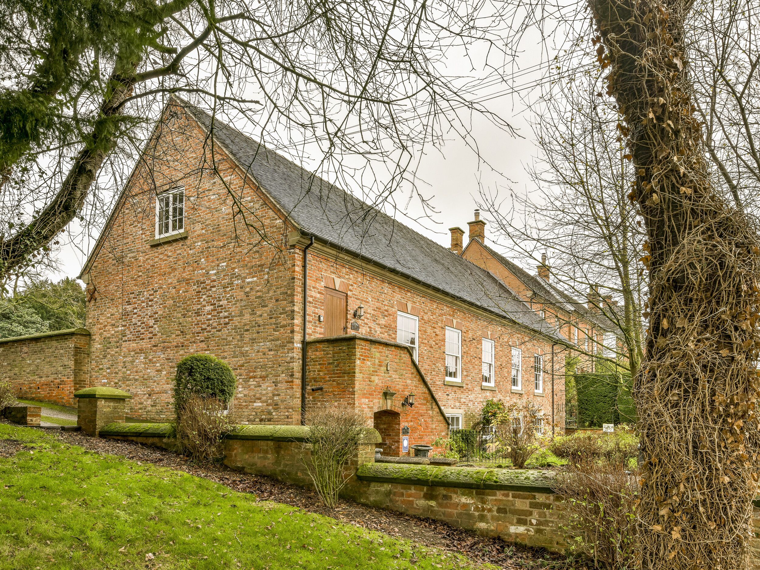 Un bâtiment traditionnel en briques, doté d’un toit à forte pente, d’une petite entrée et d’un jardin bien entretenu agrémenté d’une haie et d’un arbre.