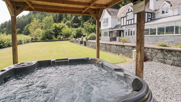 Un bain à remous sous une pergola en bois, avec un mur en pierre et une maison en arrière-plan.