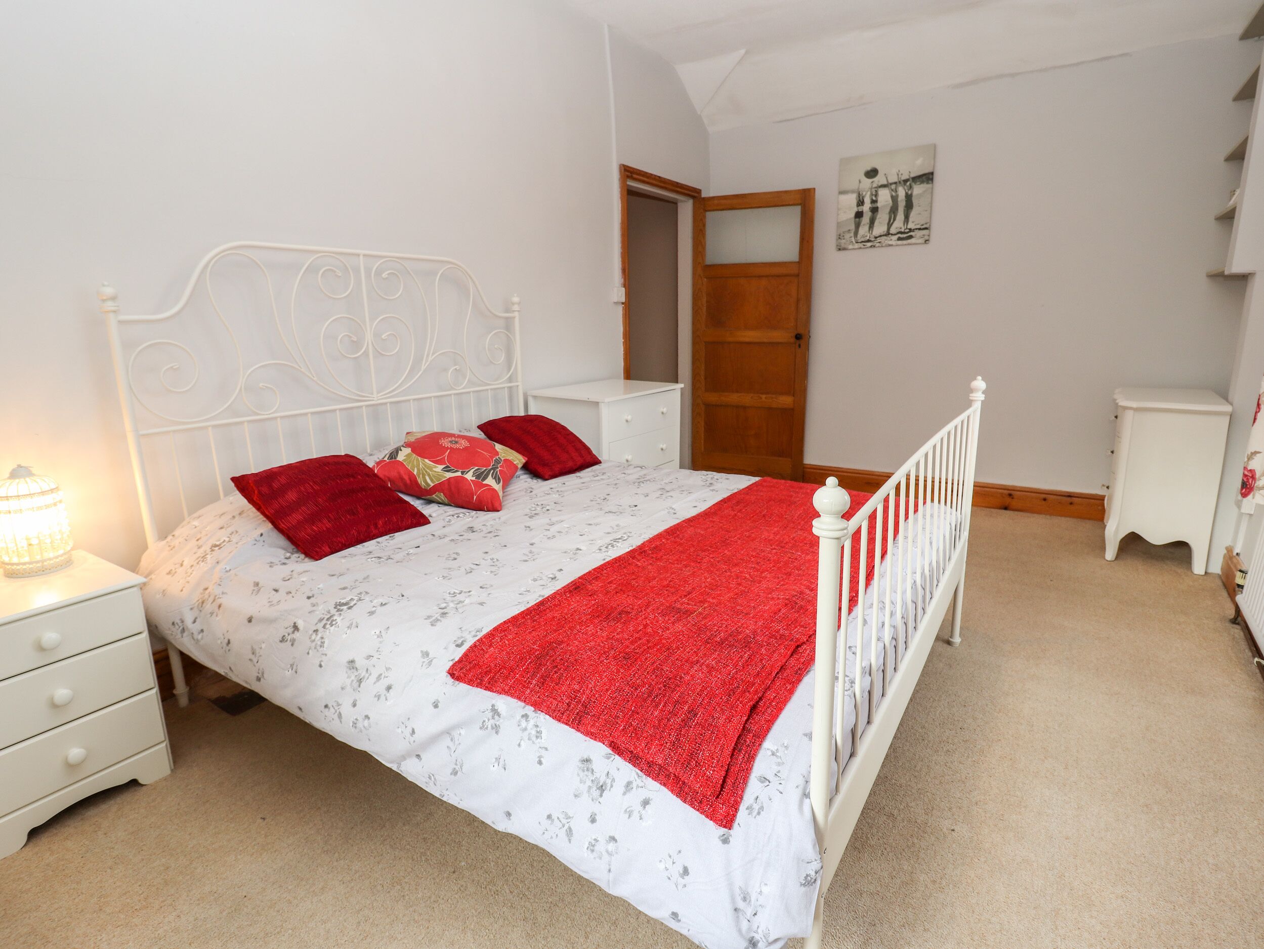 A bedroom with a white bed, red pillows, a white nightstand, a wooden door, and a small white dresser.
