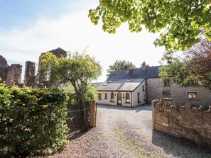 A stone building with a gravel driveway, surrounded by greenery and historical ruins. - Abbey Cottage (Denbigh)