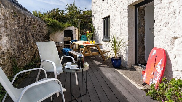 Una terraza de madera con mobiliario exterior, una tabla de surf y un muro de piedra.