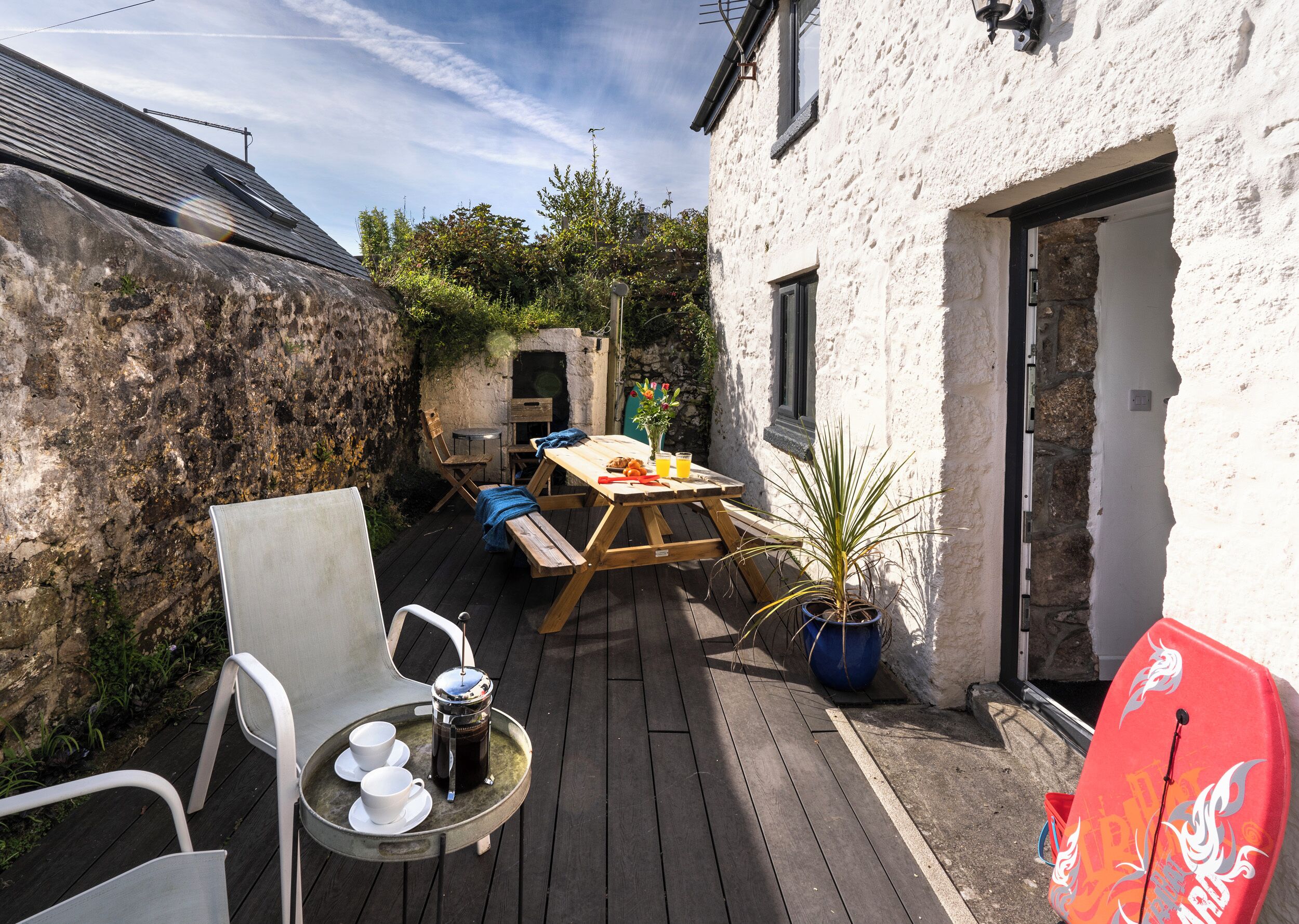 Een houten terras met een tafel, stoelen, een stenen muur en een surfplank.