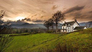 A two-story house with a white exterior, surrounded by a green field and trees. - One The Howe (Keswick)