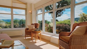 A sunlit room with wicker furniture, large windows, and a view of greenery. - One The Howe (Keswick)
