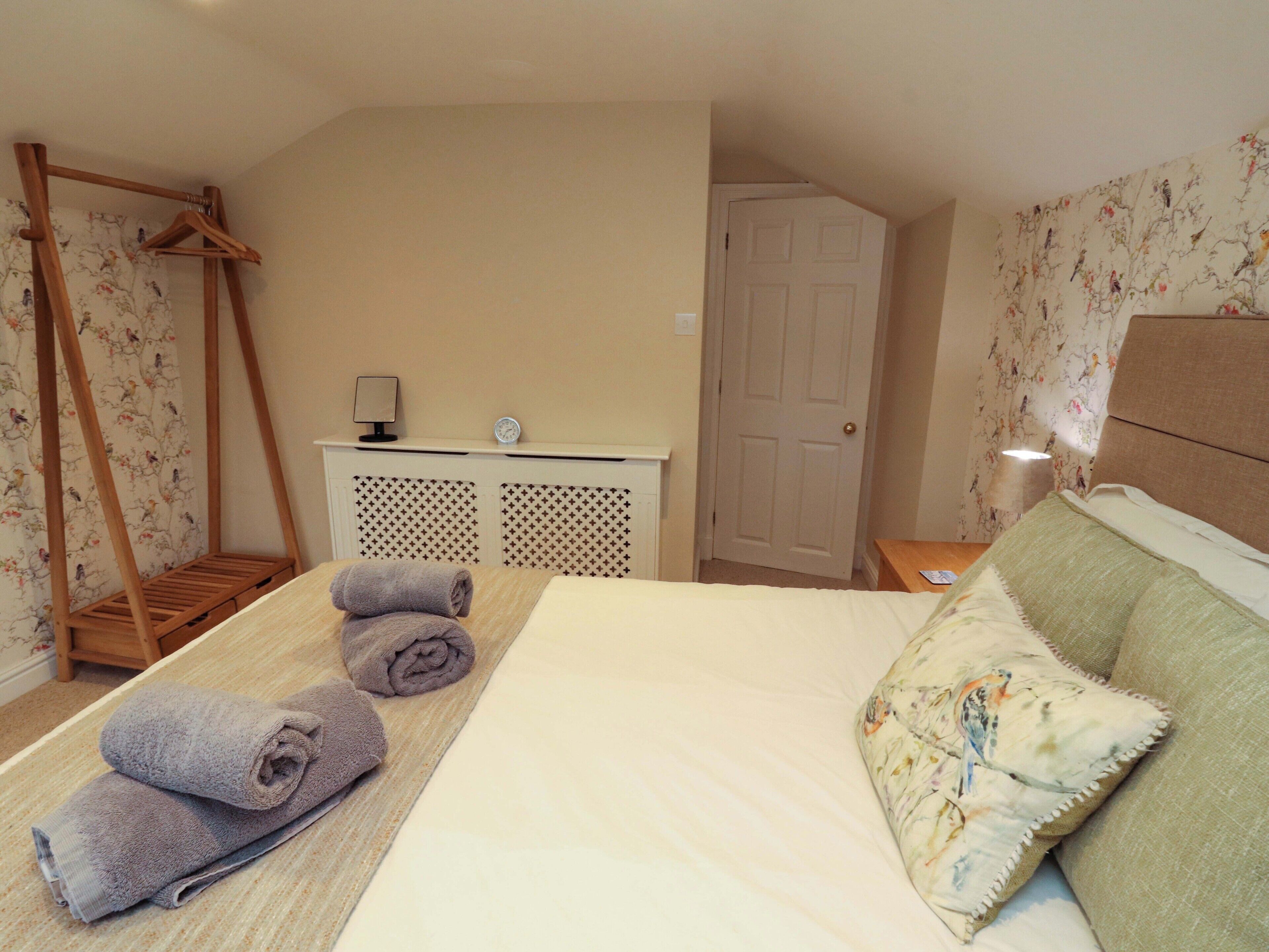A bedroom with a bed, a wooden hanger, a desk with a lamp, and a door.