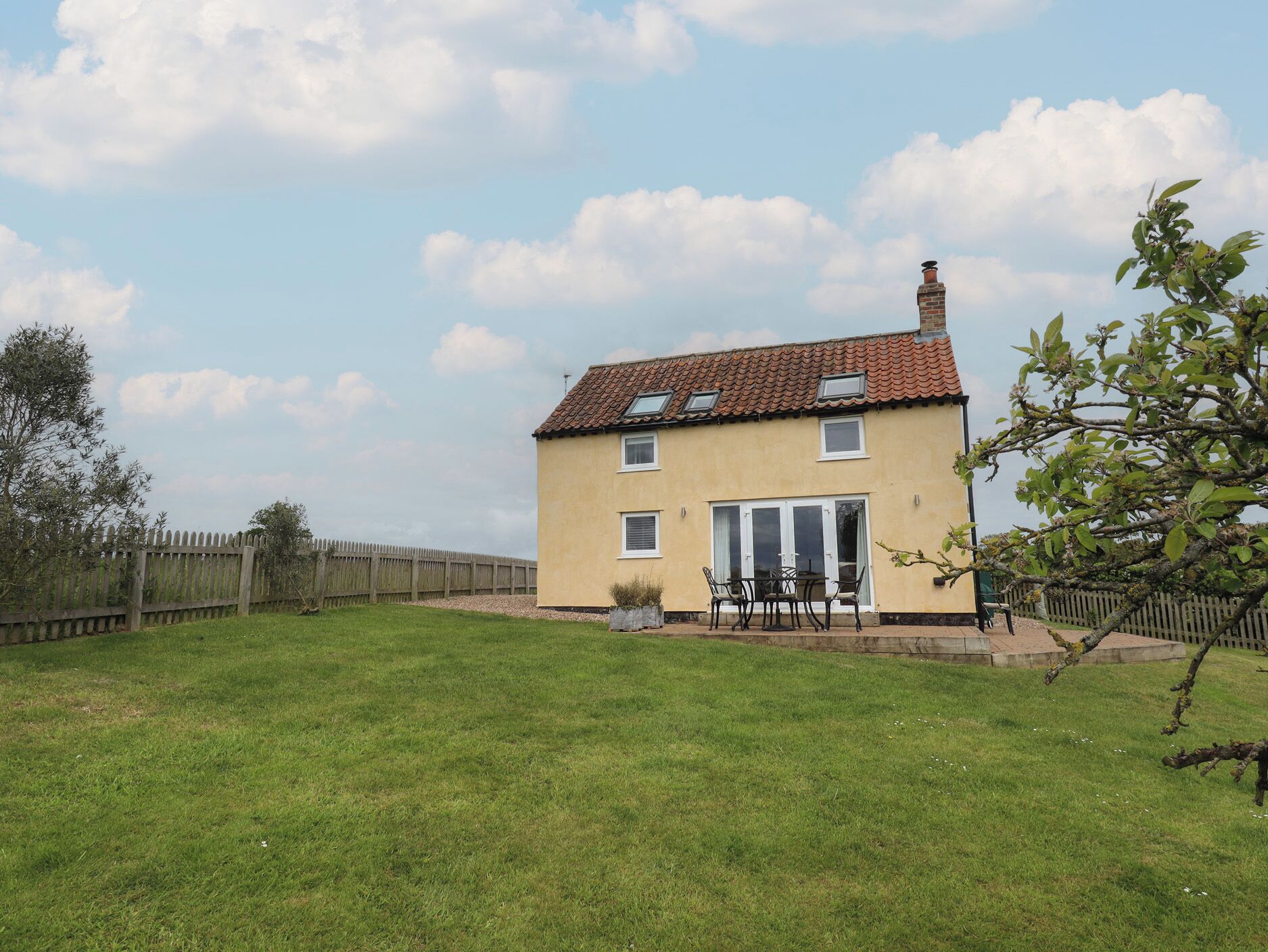 Une maison à deux étages, avec un toit en tuiles, une cour clôturée et une terrasse aménagée avec du mobilier d’extérieur.