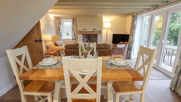A dining area with a wooden table set for four, surrounded by white chairs. There is a fireplace in the background, and a television is visible in the living area. - Shepherd's Cottage (Alford)