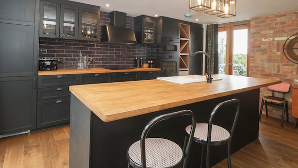 A modern kitchen with dark cabinetry, a wooden countertop, and a central island with two chairs. - The Courtyard - Hilltop Barn (Blandford Forum)
