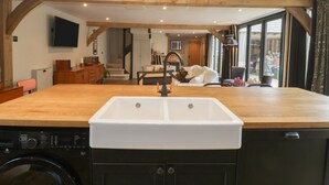 A kitchen with a large sink, wooden beams, a washing machine, and a dining area with a sofa and table. - The Courtyard - Hilltop Barn (Blandford Forum)