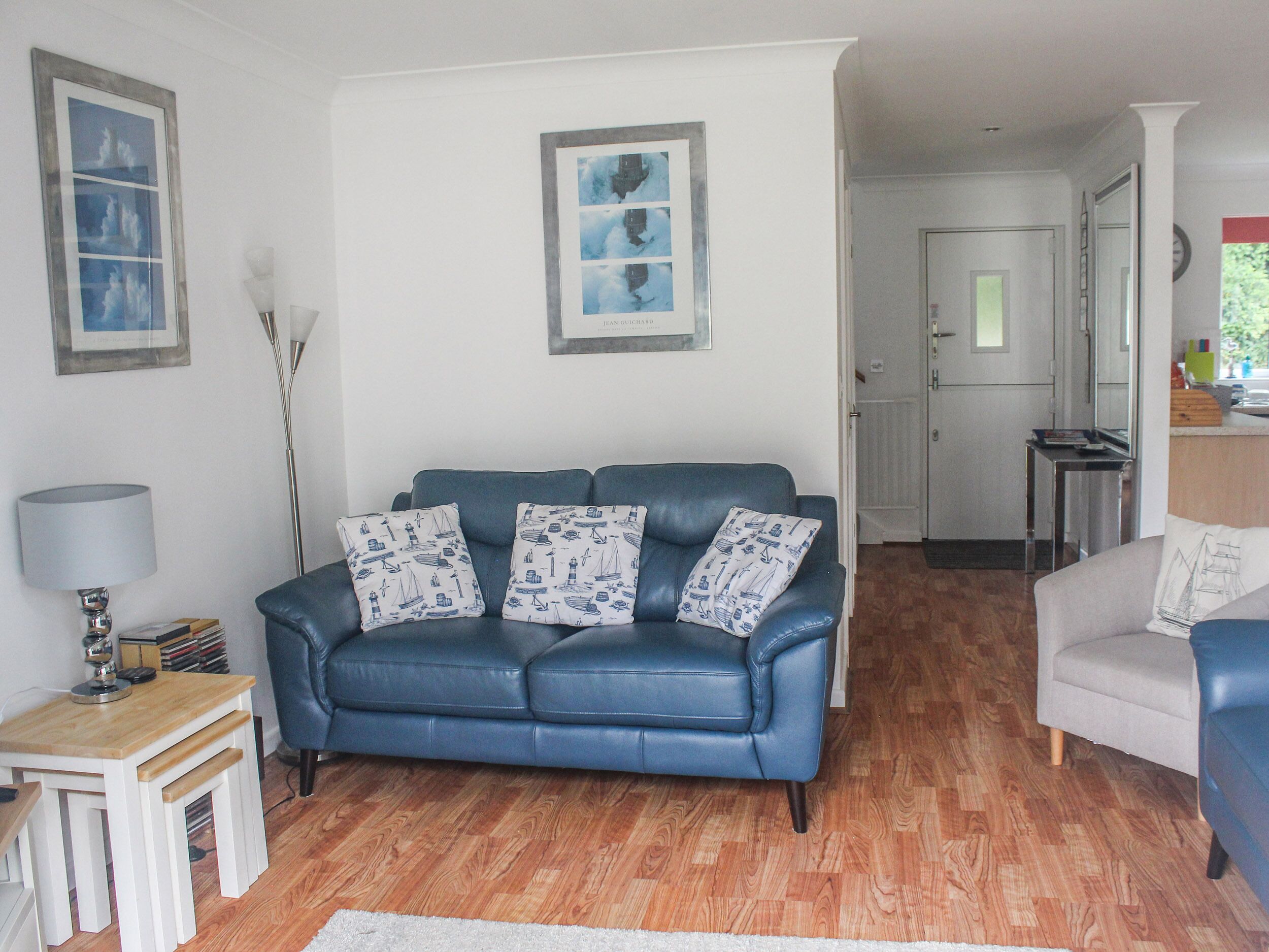 A living room with a blue sofa, a white armchair, a small wooden table, and a lamp. There are framed pictures on the wall and a door leading to another room.