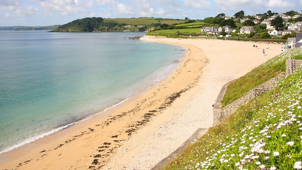 A coastal beach with a sandy shore, a clear blue sea, and a grassy hill with houses in the background. - Robinswood (Falmouth)