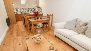 A cozy living room with a wooden dining table, a sofa, and a kitchen in the background. - Macaw Cottages No 4 (Kirkby Stephen)